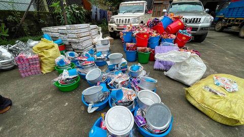 Dozens of plastic basins and buckets packed with aluminum pots, lids and mugs on a concrete yard, beside stacked cartons and bundles of folded cloth.