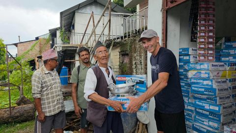 An older man in a Nepali topi and glasses takes hold of a boxed pressure cooker, a stainless steel pot and a boxed gas stove, with cartons stacked high against the wall behind him.