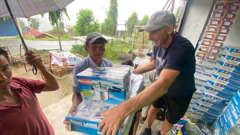Under a shelter in the rain, a man in a gray cap passes a boxed pressure cooker and a two burner gas stove to a man in a blue shirt, with stacks of the same boxes beside them.