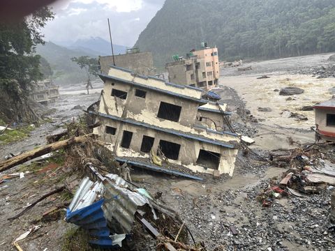 A three-story concrete building leans at a steep angle in gravel and mud, its windows empty and its floors cracked apart, with brown water running past and uprooted trees in front.