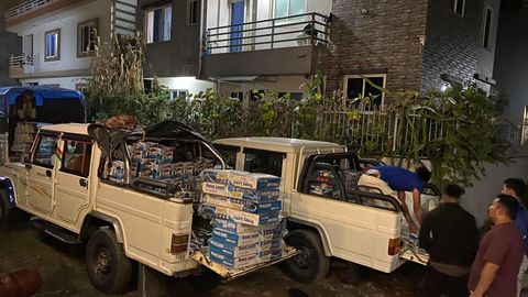 Two cream pickup trucks at night with their beds stacked with cartons of gas stoves, a man leaning over one side handling a sack.