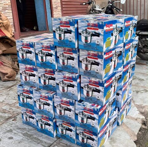 Dozens of cardboard boxes of three liter pressure cookers stacked several layers high on a pavement outside a shop with a red shutter.