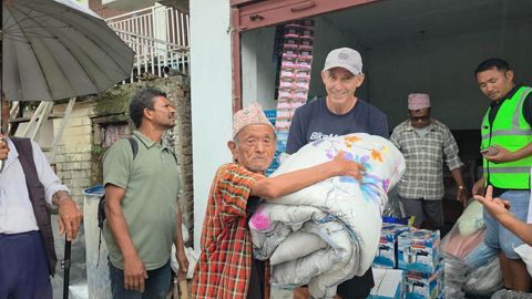 An older man in a Nepali topi and red plaid shirt holds a folded floral quilt in both arms at the front of a building stacked with relief supplies.