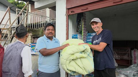 Two men hold either side of a large folded pale green quilt and pass it to an older man in a black cap, at a shop front stacked with cartons of supplies.