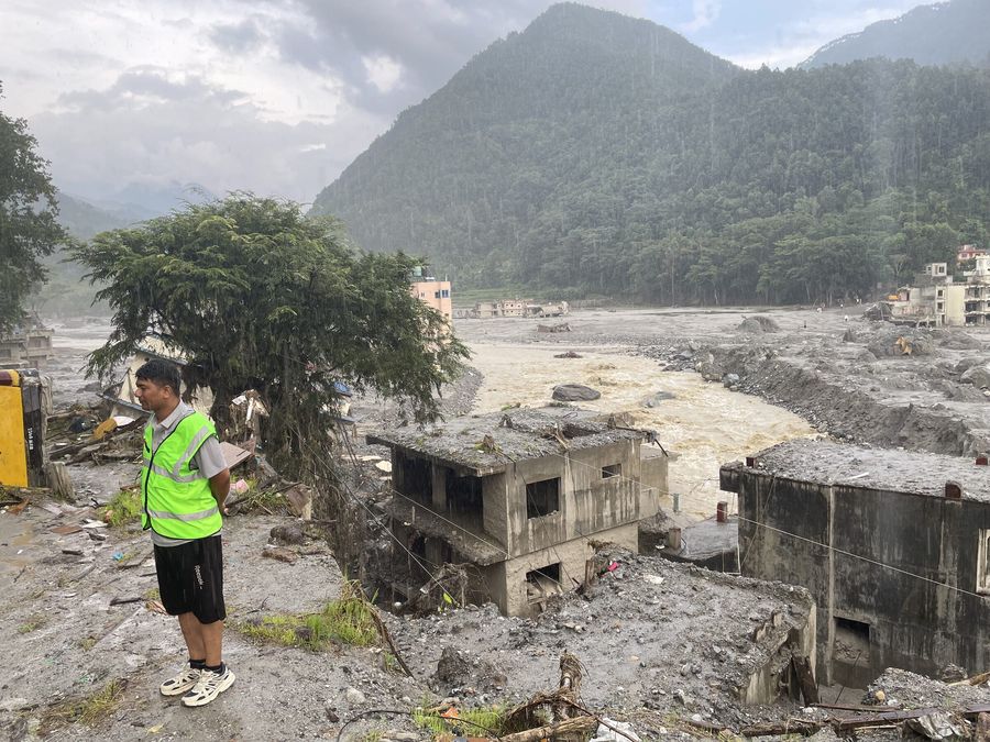 A man in a green safety vest stands on a muddy bank in the rain looking out at a fast brown river, a concrete house stripped to its frame in the silt in front of him.