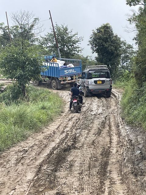 A blue flatbed truck loaded with sacks and a silver SUV climb a rutted dirt track between green trees, with a man standing beside a motorcycle.