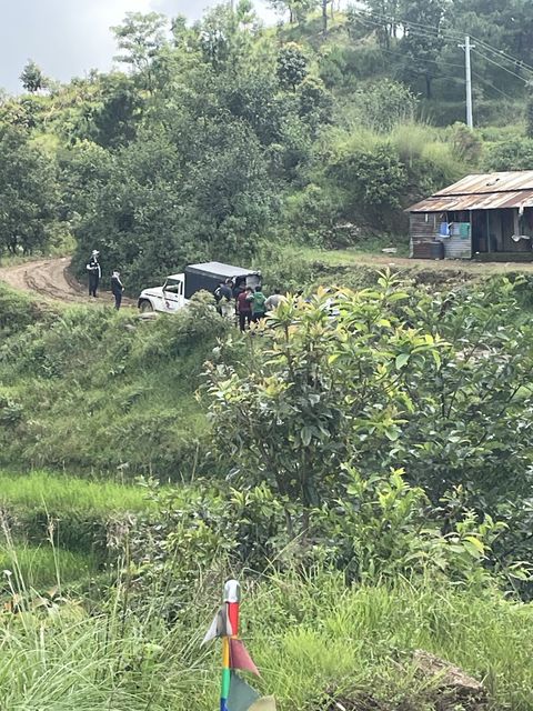 A white pickup with a dark canopy parked on a muddy hillside track, with about eight people standing at its tailgate and a tin-roofed building on the slope.