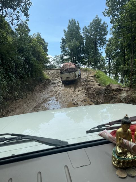 Seen through a windshield, a white pickup with a tarp-covered load climbs a steep dirt track cut into a forested hillside, its wheels in deep ruts of muddy water.