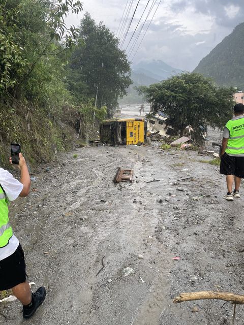 A yellow school bus lies on its side in gray mud beside a road, with a partly collapsed building and a debris-strewn riverbed behind it.