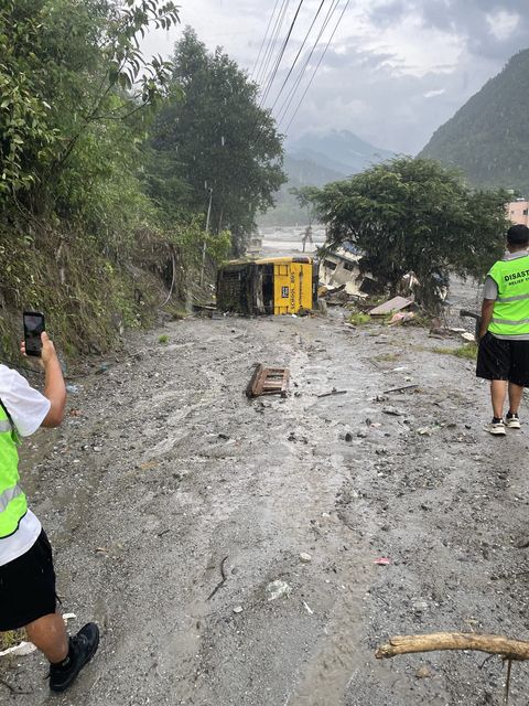 A yellow school bus lies on its side in gray mud beside a road, with a partly collapsed building and a debris-strewn riverbed behind it.