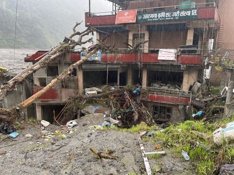A multi-story concrete building in heavy rain, its lower floors torn open and packed with mud and broken timber, with uprooted tree trunks leaning across the roof.