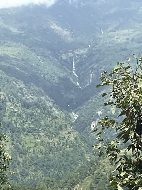 A steep green valley with terraced slopes, low cloud on the ridge, and a waterfall dropping to a pale stream on the valley floor.