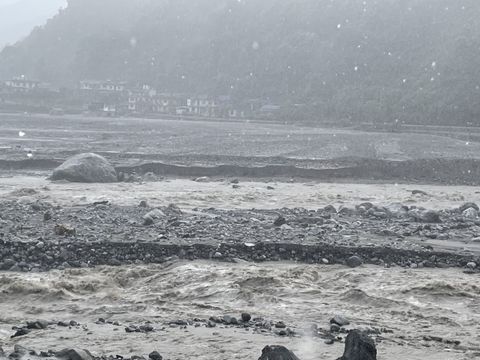 A brown river in flood fills the width of a valley, breaking over boulders, with a row of concrete houses across the water at the foot of a forested hillside.