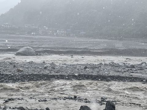 A brown river in flood fills the width of a valley, breaking over boulders, with a row of concrete houses across the water at the foot of a forested hillside.